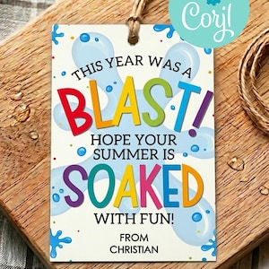 Può includere: Un'etichetta regalo rettangolare bianca con uno spago di juta, caratterizzata da una grafica a schizzi d'acqua blu e testo multicolore: THIS YEAR WAS A BLAST! HOPE YOUR SUMMER IS SOAKED WITH FUN! FROM CHRISTIAN. Un logo circolare verde acqua è nell'angolo.