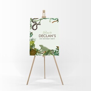 Può includere: Benvenuto al party di compleanno dell'ottavo anno di Declan a tema giungla. Il cartello presenta un'iguana verde, un serpente, una tartaruga e rane su uno sfondo verde con foglie tropicali.