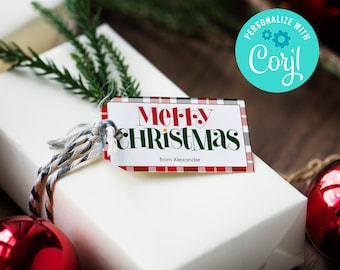 Peut inclure: Une boîte cadeau blanche avec une étiquette cadeau à carreaux rouge et vert qui dit "Joyeux Noël de la part d'Alexandre". L'étiquette est attachée à la boîte avec une ficelle. La boîte est entourée de branches de pin vertes et de boules rouges.