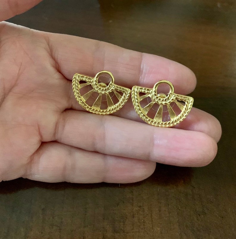 Op de afbeelding: Een paar goudkleurige oorbellen. Elke oorbel heeft een waaiervormig ontwerp met een getextureerde rand en een kleine lus aan de bovenkant. De oorbellen worden op een hand getoond.