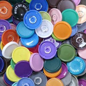 May include: A collection of colorful plastic bottle caps in various shades, including blue, green, orange, purple, and white. Some caps have text that reads "MUST BE DILUT" and "WARNING PARALYZING AGENT".