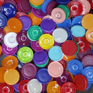 May include: A collection of colorful jar lids in various shades, including red, blue, green, yellow, orange, purple, and pink. The lids are round with a central circular design and a raised edge, creating a visually appealing assortment.