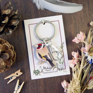 a wooden charm keyring with a swivel clasp shaped and illustrated as a goldfinch bird displayed on a branded backing card
