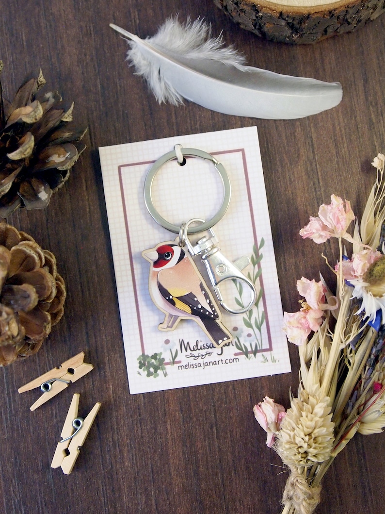 a wooden charm keyring with a swivel clasp shaped and illustrated as a goldfinch bird displayed on a branded backing card