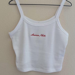 May include: White crop top with thin straps, hanging on a black wire hanger. The text "American Made" is embroidered in red across the chest. The top is made of a ribbed fabric.