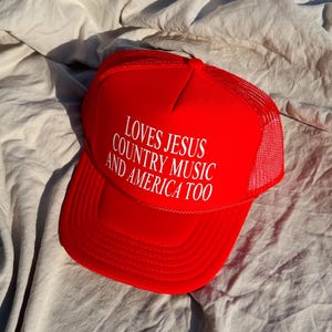 May include: A red trucker hat with the text "LOVES JESUS COUNTRY MUSIC AND AMERICA TOO" in white. The hat features a mesh back and a curved peak. It rests on a wrinkled, light-coloured fabric.