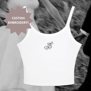 Może przedstawiać: Biały top na ramiączkach z cienkimi ramiączkami i małym haftowanym wzorem dwóch serc i kokardy. Tekst "CUSTOM EMBROIDERY!" znajduje się na grafice w kształcie gwiazdy. Obraz jest czarno-biały.