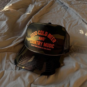 May include: Camouflage trucker hat with the text "GOOD COLD BEER AND COUNTRY MUSIC" in orange. The hat has a black mesh back and a curved brim. The text "THE ONLY THINGS KEEPING ME FROM LOSING IT" is below the main text.