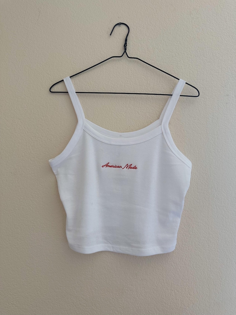 May include: White crop top with thin straps, hanging on a black wire hanger. The text "American Made" is embroidered in red across the chest. The top is made of a ribbed fabric.
