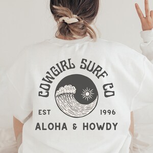 Może przedstawiać: Biała koszulka z czarno-białym nadrukiem fali i słońca. Na koszulce widnieje napis "Cowgirl Surf Co. Est 1996 Aloha & Howdy".