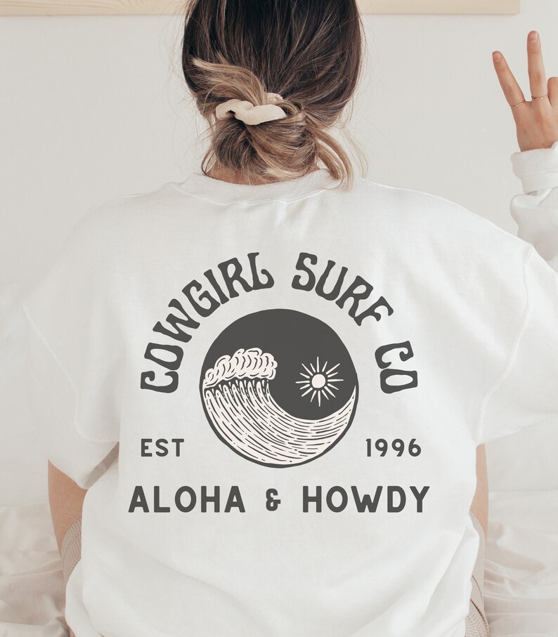 Może przedstawiać: Biała koszulka z czarno-białym nadrukiem fali i słońca. Na koszulce widnieje napis "Cowgirl Surf Co. Est 1996 Aloha & Howdy".