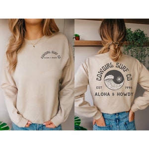 Może przedstawiać: Beżowa bluza z czarnym nadrukiem graficznym z przodu i z tyłu. Nadruk graficzny zawiera napis "Cowgirl Surf Co. Aloha & Howdy" oraz falę i słońce.