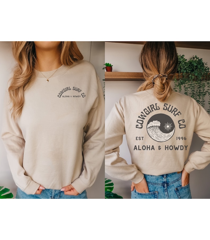 Może przedstawiać: Beżowa bluza z czarnym nadrukiem graficznym z przodu i z tyłu. Nadruk graficzny zawiera napis "Cowgirl Surf Co. Aloha & Howdy" oraz falę i słońce.
