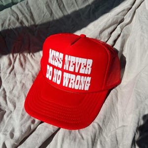 May include: Red trucker hat with a white mesh back. The front of the hat has white text that reads "Miss Never Do No Wrong."