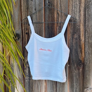 May include: White tank top with thin straps, hanging on a black hanger against a wooden background. The text "American Made" is embroidered in red across the chest. The top is made of a soft, lightweight material.