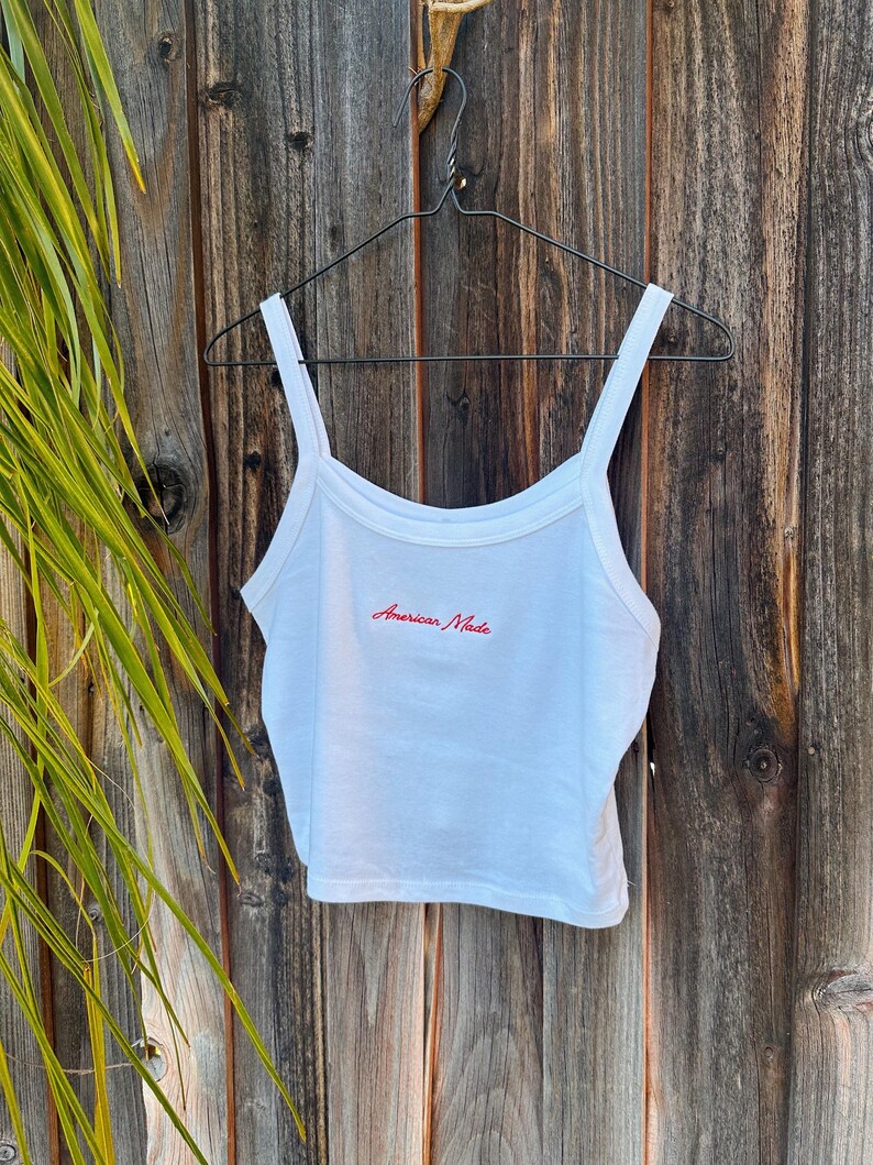 May include: White tank top with thin straps, hanging on a black hanger against a wooden background. The text "American Made" is embroidered in red across the chest. The top is made of a soft, lightweight material.