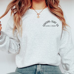 Może przedstawiać: Szara bluza z nadrukiem "Cowgirl Surf Co. Aloha & Howdy" na przodzie.
