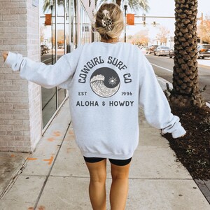Może przedstawiać: Szara bluza z czarno-białym nadrukiem fali i słońca. Tekst na bluzie brzmi "Cowgirl Surf Co. Est. 1996 Aloha & Howdy".