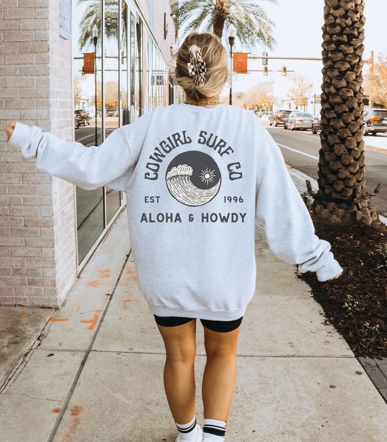 Może przedstawiać: Szara bluza z czarno-białym nadrukiem fali i słońca. Tekst na bluzie brzmi "Cowgirl Surf Co. Est. 1996 Aloha & Howdy".