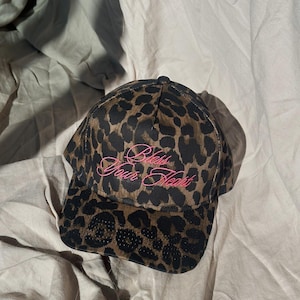 May include: A baseball cap featuring a brown and black leopard print pattern. The front of the cap displays the embroidered text "Bless Your Heart" in a bright pink cursive font. The cap has a structured crown and a curved brim.