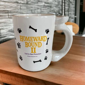 May include: White ceramic mug with the text "HOMEWARD BOUND II" in yellow, along with black paw prints and bone graphics. The mug has a unique handle design and the text "Lost in San Francisco".