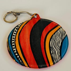 May include: A round keychain with a colorful abstract design. The keychain features a silver metal ring and a leather pendant with a pattern of orange, black, yellow, blue, and white stripes and dots. The pendant is approximately 3 inches in diameter.