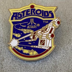 May include: A vintage Atari Asteroids pin. The pin features a shield shape with the word "Asteroids" in blue and gold lettering. The Atari logo is also visible. The pin has a white and red spaceship design shooting a white laser beam.