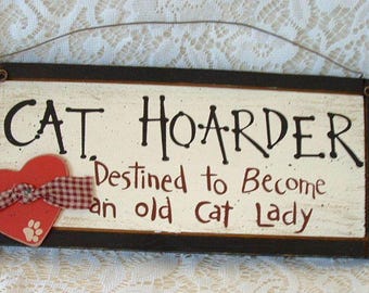 May include: Wooden sign with the words "CAT HOARDER" in black, with the phrase "Destined to Become an old Cat Lady" in red. A red heart with a paw print and a plaid ribbon is attached to the left side.