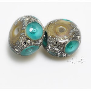 Op de afbeelding: Twee ronde glazen kralen met een bruin, zilver en turquoise kleurenschema. De kralen hebben een gespikkeld patroon en een gat in het midden.