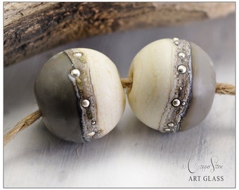 May include: Two round glass beads with a white and gray color scheme. The beads have a silver metallic band with small silver dots around the band. The beads are strung on a natural fiber cord.