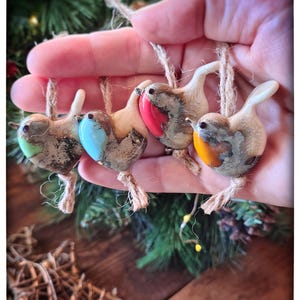 Op de afbeelding: Vier handgemaakte vogelornamenten met jute touwhangers. Elke vogel heeft een uniek kleurenschema: groen, blauw, rood en oranje. De ornamenten zijn gemaakt van een gespikkeld, getextureerd materiaal. Een hand houdt de vogels vast, met een kerstboom op de achtergrond.
