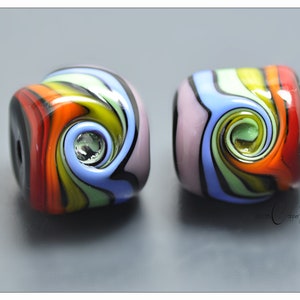 Puede incluir: Dos cuentas de vidrio cuadradas con un patrón de remolinos en colores rojo, naranja, amarillo, verde claro, azul claro, lavanda y negro. Cada cuenta tiene un orificio central y un acabado brillante. El texto Copper Stone es visible en la esquina inferior derecha.