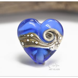 Può includere: Un ciondolo a forma di cuore in vetro blu con un design a spirale e dettagli argentati e marroni. Il ciondolo è fatto a mano e ha una finitura lucida.