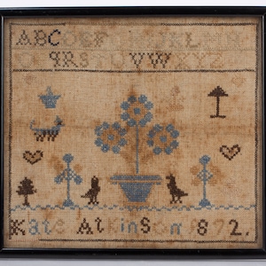 May include: A framed sampler with blue and brown embroidery depicting a tree, birds, and a heart. The sampler includes the letters ABC and the text "Done at 182."