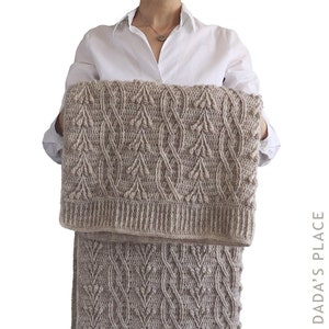 May include: A beige crocheted blanket with a leaf pattern. The blanket is folded in half and held by a person.