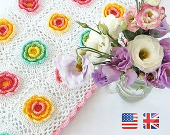 Puede incluir: Una manta de bebé de crochet blanco con un borde festoneado y motivos de flores de colores. La manta está hecha con un delicado patrón de puntadas y presenta una variedad de colores pastel, incluyendo rosa, amarillo, verde y azul. La manta se muestra sobre una superficie blanca con un ramo de flores en un frasco de vidrio.