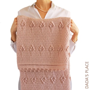 May include: A light pink crocheted blanket with a textured pattern and leaf-like designs. The blanket is held up, showcasing its detailed stitching and soft colour. The blanket has a decorative border and is labeled with the text "DADA'S PLACE".