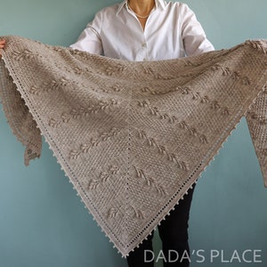 May include: A beige crocheted shawl with a delicate leaf pattern and beaded edging. The shawl is held by a person wearing a white shirt and black pants.