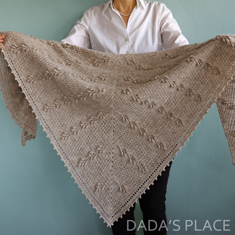 May include: A beige crocheted shawl with a delicate leaf pattern and beaded edging. The shawl is held by a person wearing a white shirt and black pants.