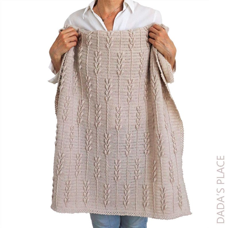 Puede incluir: Una manta de ganchillo beige con un patr&oacute;n vertical repetitivo de puntos en relieve en forma de hoja, separados por columnas de puntos altos. El tejido tiene una superficie texturizada y un color tostado claro uniforme en toda la pieza.
