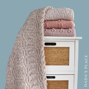 May include: A stack of three knitted blankets in shades of pink and beige, with a fourth blanket draped over the side. The blankets are on a white bedside table with woven basket drawers. The draped blanket has a leaf pattern. Text on the right side reads "DADA'S PLACE".