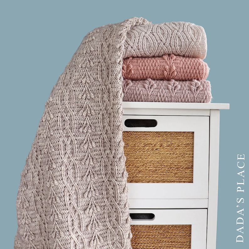 May include: A stack of three knitted blankets in shades of pink and beige, with a fourth blanket draped over the side. The blankets are on a white bedside table with woven basket drawers. The draped blanket has a leaf pattern. Text on the right side reads "DADA'S PLACE".