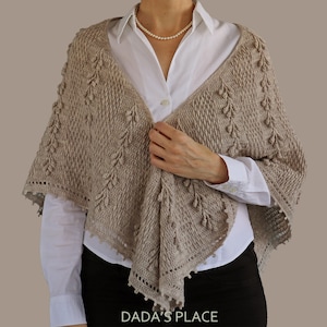 May include: A beige crochet shawl with a delicate lace trim and a textured pattern. The shawl is draped over a person's shoulders and is held in place by their hand.