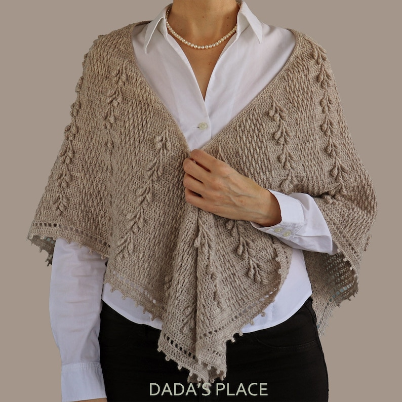 May include: A beige crochet shawl with a delicate lace trim and a textured pattern. The shawl is draped over a person's shoulders and is held in place by their hand.