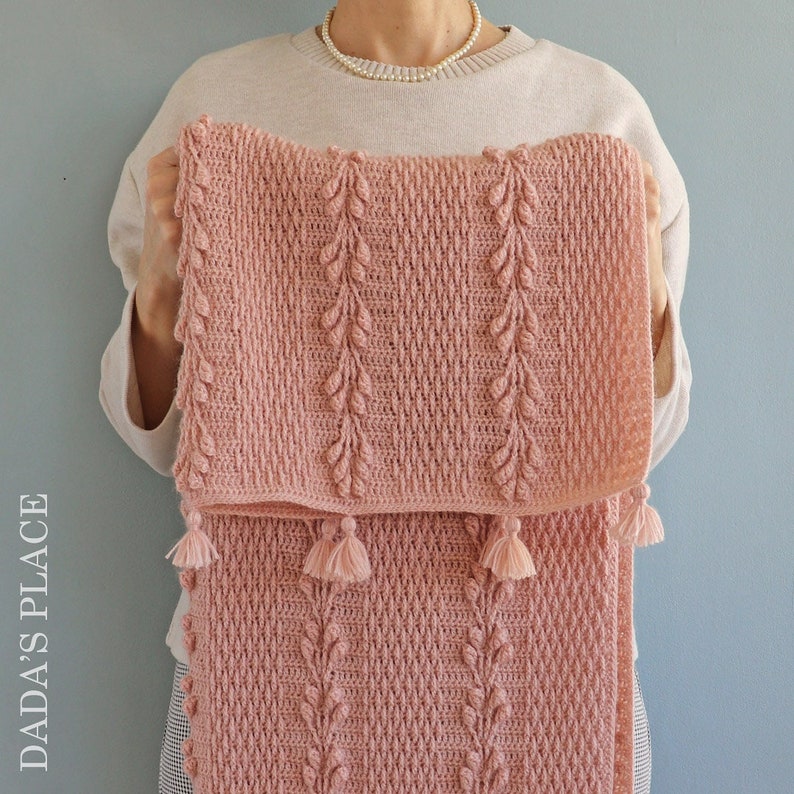 May include: A pink crocheted blanket with a leaf pattern and tassels. The blanket is folded in half and held by a person.