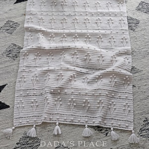May include: A white crocheted throw blanket with a delicate floral pattern and tassels along the bottom edge. The blanket is folded and lying on a patterned rug.