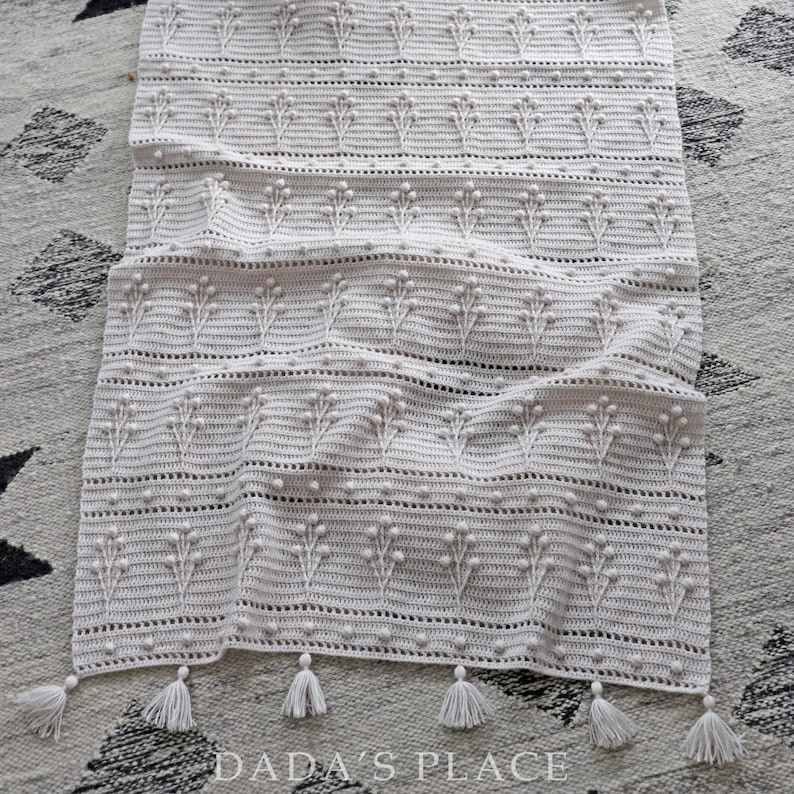 May include: A white crocheted throw blanket with a delicate floral pattern and tassels along the bottom edge. The blanket is folded and lying on a patterned rug.