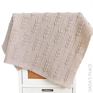 Puede incluir: Una manta de ganchillo beige con un patr&oacute;n vertical repetitivo de puntos en relieve en forma de hoja, separados por columnas de puntos altos. La manta tiene un borde decorativo y el texto DADA'S PLACE es visible en la esquina inferior derecha.
