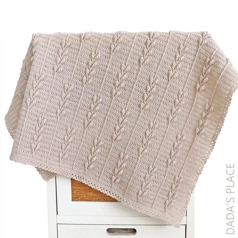 Puede incluir: Una manta de ganchillo beige con un patr&oacute;n vertical repetitivo de puntos en relieve en forma de hoja, separados por columnas de puntos altos. La manta tiene un borde decorativo y el texto DADA'S PLACE es visible en la esquina inferior derecha.