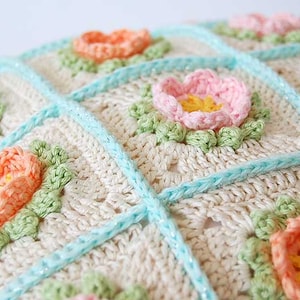 May include: A crocheted pillow with a white background and a pattern of pastel pink, peach, and yellow flowers. The pillow is trimmed with a light blue border.